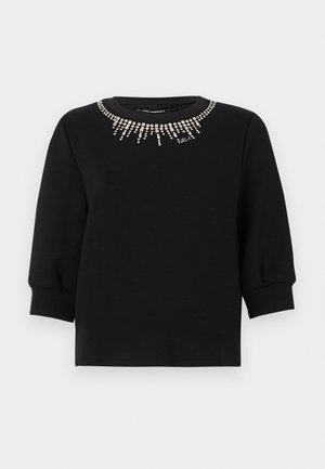 Crna majica s tri četvrt rukavima i okruglim izrezom na vratu ukrašenim polukružnim uzorkom od srebrnih rhinestona i malim potpisnim detaljem.