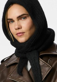 Sciarpa nera a maglia con un tessuto testurizzato, avvolta strettamente attorno al collo, abbinata a una giacca di pelle marrone con zip dotata di risvolti prominenti.