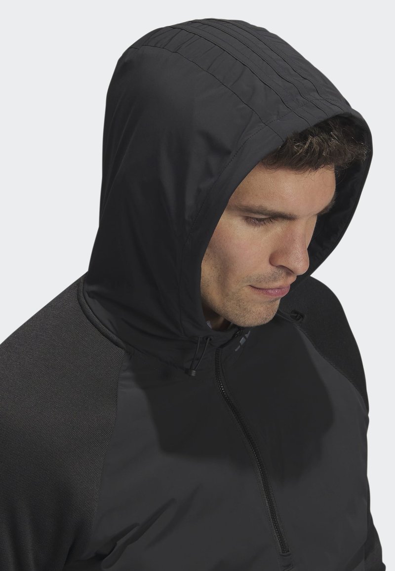 Mann trägt eine schwarze Kapuzenjacke mit Reißverschluss und schaut nach unten, vor einem schlichten hellgrauen Hintergrund.