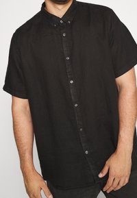Camicia nera a maniche corte con bottoni, realizzata in tessuto leggero, con colletto classico, bottoni frontali e una vestibilità morbida.