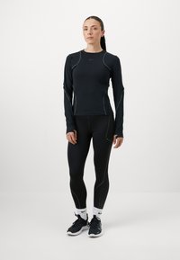 Zwarte sporttop met lange mouwen en contrasterende naden, gecombineerd met zwarte leggings met geperforeerde accenten en zwarte hardloopschoenen.