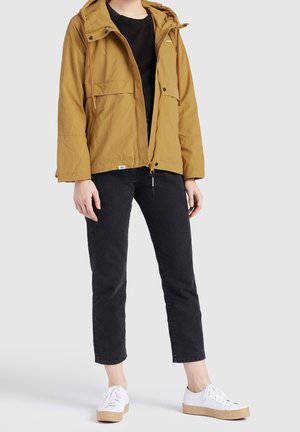 Osoba ubrana w musztardową żółtą kurtkę z kapturem, czarne krótkie spodnie i białe trampki, stojąca na jasnoszarym tle.