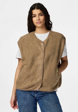 Jonge vrouw draagt een beige, gestructureerd vest met knopen over een wit T-shirt met korte mouwen en een blauwe spijkerbroek, staand met één hand in de zak.