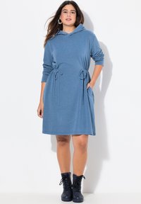 Kapuzenkleid aus blauer Baumwollmischung, mit Seitentaschen, Kordelzug in der Taille und langen Ärmeln. Kombiniert mit schwarzen Schnürboots.