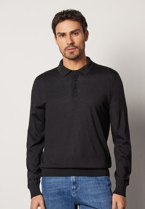 Mann mit kurzen dunklen Haaren und Bart, der ein schwarzes Langarm-Poloshirt und blaue Jeans trägt, steht vor neutralem Hintergrund.