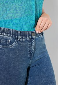 Jeans i denim med en medelblå tvätt, med en slät yta, tryckknappsstängning och en lätt stretch, bärs tillsammans med en blå mönstrad topp.