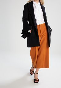 Sort skræddersyet frakke med flagrende ærmer, hvid bluse med flæser, orange vidde bukser og sorte sandaler med blomster detaljer.