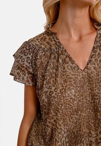 Blusa con stampa animale marrone, con maniche corte a volant e scollo a V. Il tessuto presenta un design plissettato e testurizzato con una leggera lucentezza.