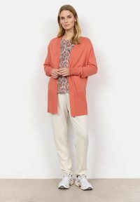 Korallfärgad stickad cardigan med öppen framsida, långa ärmar, matchad med en mönstrad topp och krämfärgade byxor, bärs med ljusa sneakers.