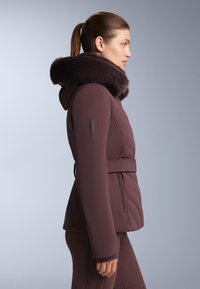 Uskladišena bordo jakna od punjene tkanine s tamnim, mekanim krznenim kapuljačom, strukirana pojasom i prošivenim uzorkom, uparena s odgovarajućim uskim hlačama.