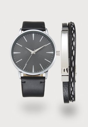 Montre ronde noire avec des index et aiguilles argentés, associée à deux bracelets en cuir noir, dont un tressé avec un fil blanc.