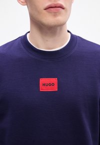 Tengerészi crewneck pulóver pamut keverékből, fekete "HUGO" felirattal ellátott piros négyzetes folttal. Bordás nyak és texturált anyag jellemzi.