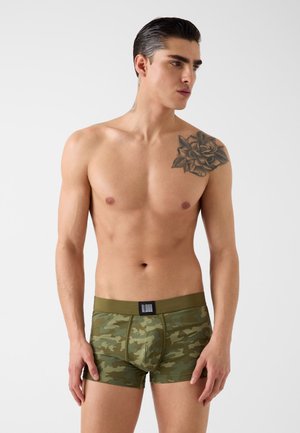 Hemdloser Mann mit floraler Schultertattoo, trägt grüne Camouflage-Boxershorts, steht vor einem schlichten hellen Hintergrund.
