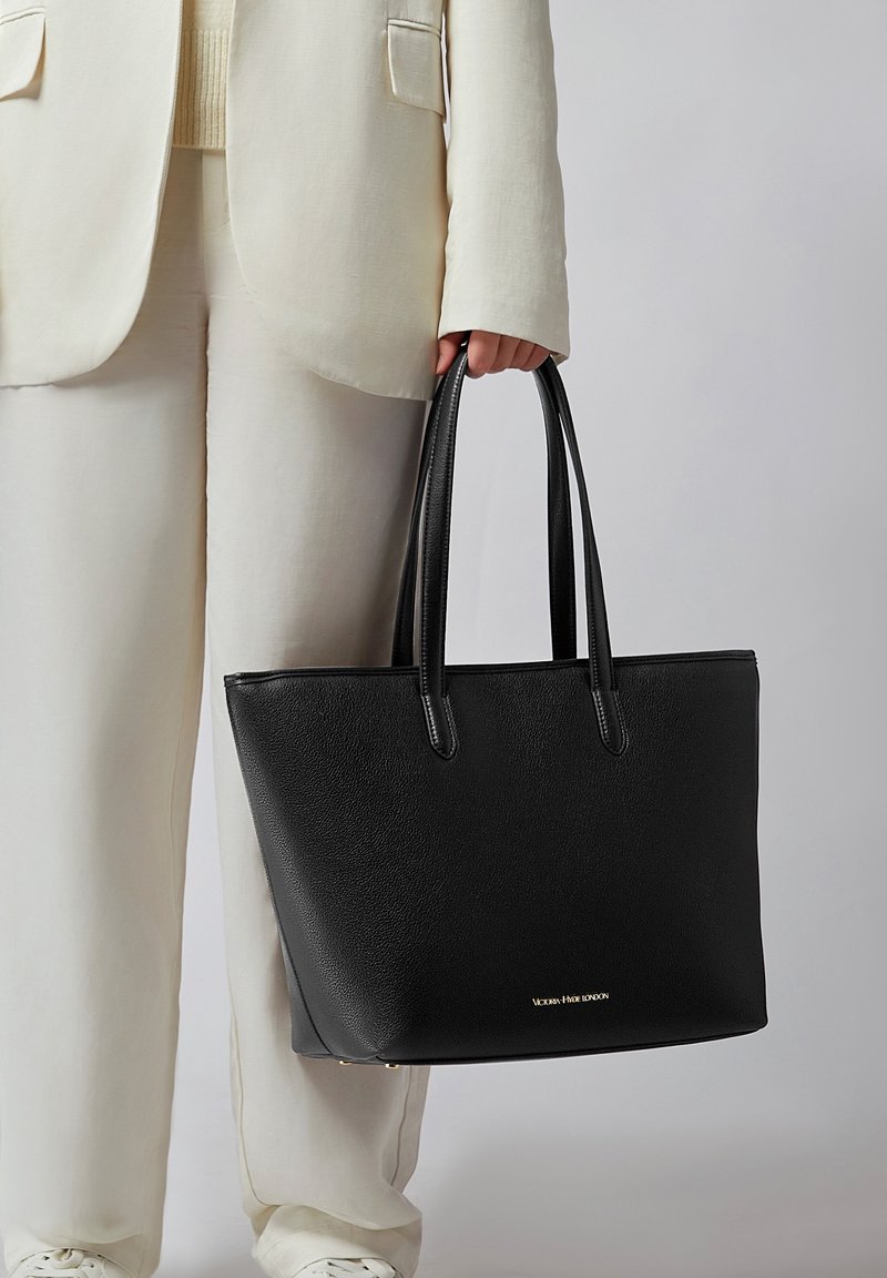 Personne en costume crème tenant un grand sac fourre-tout en cuir noir avec doubles poignées et logo doré en bas au centre.