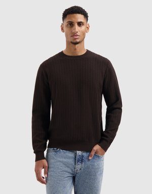 Bruine gebreide trui met een textuurpatroon, met ribbelmanchetten en -zoom, gedragen over lichtblauwe spijkerbroek. Model staat met handen in de zakken.