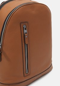 Sac à dos en cuir tan avec une forme arrondie, doté d'une fermeture éclair proéminente, d'une poche frontale et d'une surface texturée. Logo en bas.