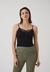 Débardeur noir avec une bordure en dentelle le long du décolleté. Bretelles spaghetti réglables. Porté avec un pantalon texturé vert olive. Design simple et ajusté.