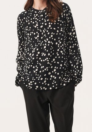 Black blouse with white polka dots, round neckline, and long sleeves. Features button placket and loose, flowing fit. Paired with black trousers.