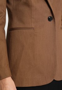 Blazer marrone realizzato in tessuto texture, caratterizzato da un solo bottone nero e una tasca a filetto, con una vestibilità sartoriale e un design strutturato.