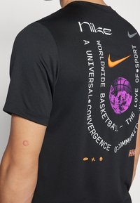 Černé tričko Nike s oranžovým swooshem, fialovou grafikou basketbalového míče a bílým zakřiveným textem o basketbalové komunitě a konvergenci.