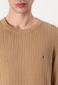 Pull beige en tricot avec une texture côtelée, col rond et un petit logo sur la poitrine. Vue en gros plan.