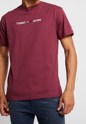 Homme portant un t-shirt Tommy Jeans bordeaux et un jean bleu foncé, debout avec les bras détendus le long du corps.