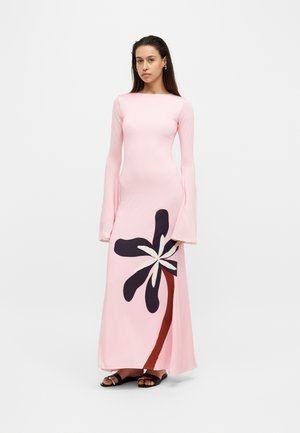 Femme portant une longue robe rose avec des manches évasées et un grand motif floral noir et blanc près de l'ourlet, debout devant un fond uni.