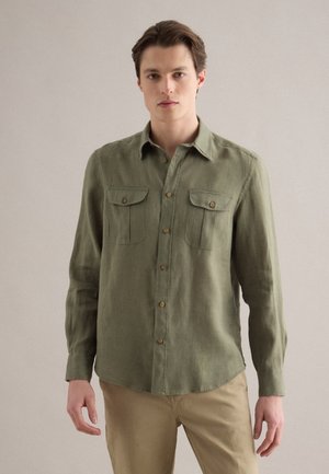 Junger Mann trägt ein olivgrünes, langärmliges Hemd mit Knöpfen und Brusttaschen sowie beige Hose, steht vor einem einfarbigen Hintergrund.