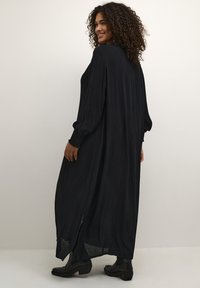 Long black dress with a flowing design, pleated texture, and puffed sleeves. Features side slits and paired with ankle boots.