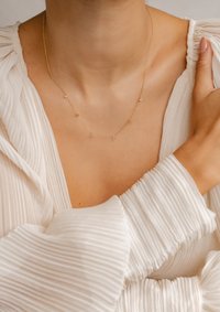 Collana in oro con piccoli accenti decorativi e pietre trasparenti, indossata intorno al collo sopra un indumento bianco testurizzato.