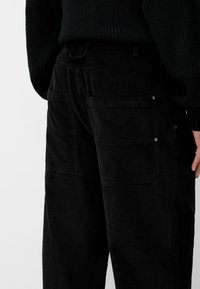 Black corduroy pants featuring a wide-leg design, textured fabric, multiple side pockets, and metal hardware accents on the pockets.