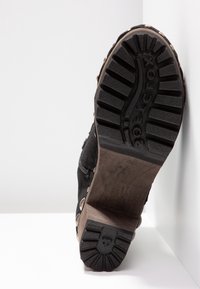 Semelle d'une botte noire à talons hauts avec des semelles profondes, des clous en métal sur le côté et un talon bloc, photographiée sur un fond blanc.