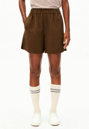 Personne portant un short marron à taille élastique, des chaussettes blanches montantes à rayures, des chaussures blanches sans lacets, et un haut rayé avec les mains dans les poches.