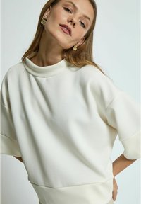 Biała bluza z wysokim kołnierzem, z oversize'owymi krótkimi rękawami, gładką fakturą i luźnym krojem. Modelka ma na sobie złote kwiatowe kolczyki.