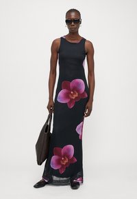Robe maxi en tissu noir avec de grands imprimés d'orchidées roses et violettes, design sans manches et silhouette ajustée, associée à un grand sac noir.