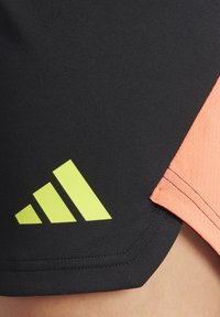 Zwarte sportshorts met een koraalkleurige accent, voorzien van een groot geel Adidas-logo. De materialen lijken glad, met een licht gestructureerde afwerking.