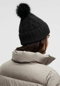 Gorro de lana negro con un patrón texturizado, con un pompón esponjoso en la parte superior y un dobladillo de canalé. Llevado sobre una chaqueta acolchada de color beige claro.