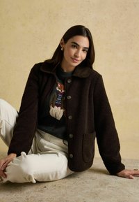 Hnedý flísový kabát s oblejším golierom, prednými gombíkmi a bočnými vreckami, nosený cez vzorovaný tmavý sveter a svetlé nohavice.