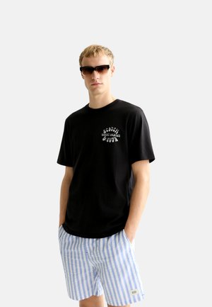 Mann trägt schwarzes Logo-T-Shirt, blau-weiß gestreifte Shorts und dunkle Sonnenbrille, steht mit Händen in den Taschen vor weißem Hintergrund.