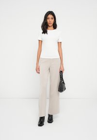 Femme portant un haut blanc à manches courtes, un pantalon large beige, des bottes à talons noires, tenant un sac à main noir à motifs sur un fond blanc.