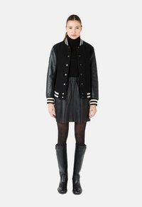 Zwarte varsity jacket met leren mouwen, crèmekleurige strepen op de manchetten, gecombineerd met een zwarte leren rok en kniehoge zwarte laarzen.