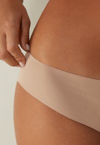 Beige naadloze onderkleding met een gladde textuur en een brede tailleband. Hand die de tailleband aanpast op een gebruinde huidkleur.
