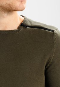 Maglione verde oliva realizzato in morbido filato, con un particolare dettaglio zip sulla spalla e sottili accenti a costine sul collo.