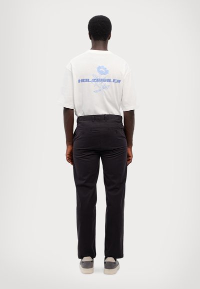 White short-sleeve t-shirt featuring blue floral graphic and text on back, paired with black trousers and gray sneakers. Minimalist design.