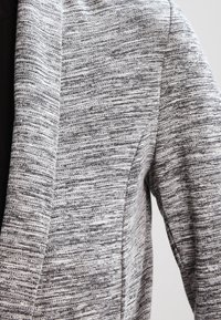 Šedý melírovaný pletený blejzr s texturovanou tkaninou, klopovým límcem s výstřihem a čistými liniemi. Má volnější střih a jemné šicí detaily.