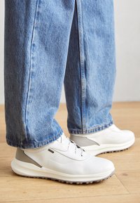 Baskets blanches en matière texturée avec des accents gris, associées à un jean en denim bleu ample. Semelle en caoutchouc et détails de laçage visibles.