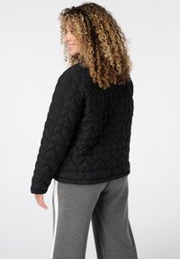 Vrouw met krullend haar, gekleed in een zwarte gewatteerde jas met hartpatronen en grijze broek met witte zijstrepen, staand in zijaanzicht.