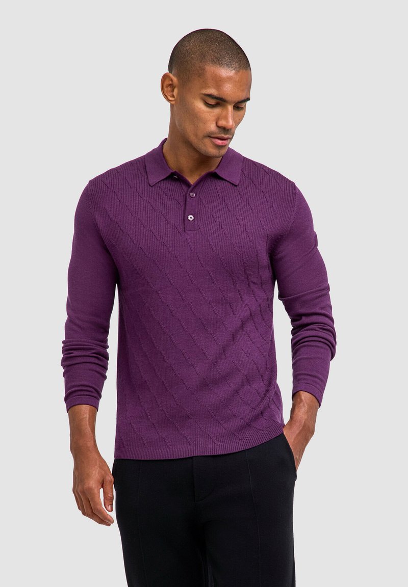 Paarse polo met lange mouwen, voorzien van een gestructureerd ruitpatroon, een placket met twee knopen en ribgebreide manchetten. Gepaard met zwarte broek.