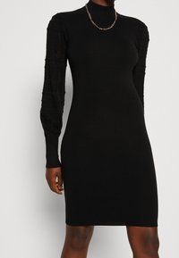 Sort kjole med en figursyet silhuet, der har lange, strukturerede blondesleeves og en høj hals. Glat tekstur med et ribbet design.