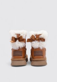 Botines marrones de cuero con ribete de piel blanca, detalles de cinta en la parte trasera y suelas de goma texturizadas. Incluye un parche con el logo en el talón.